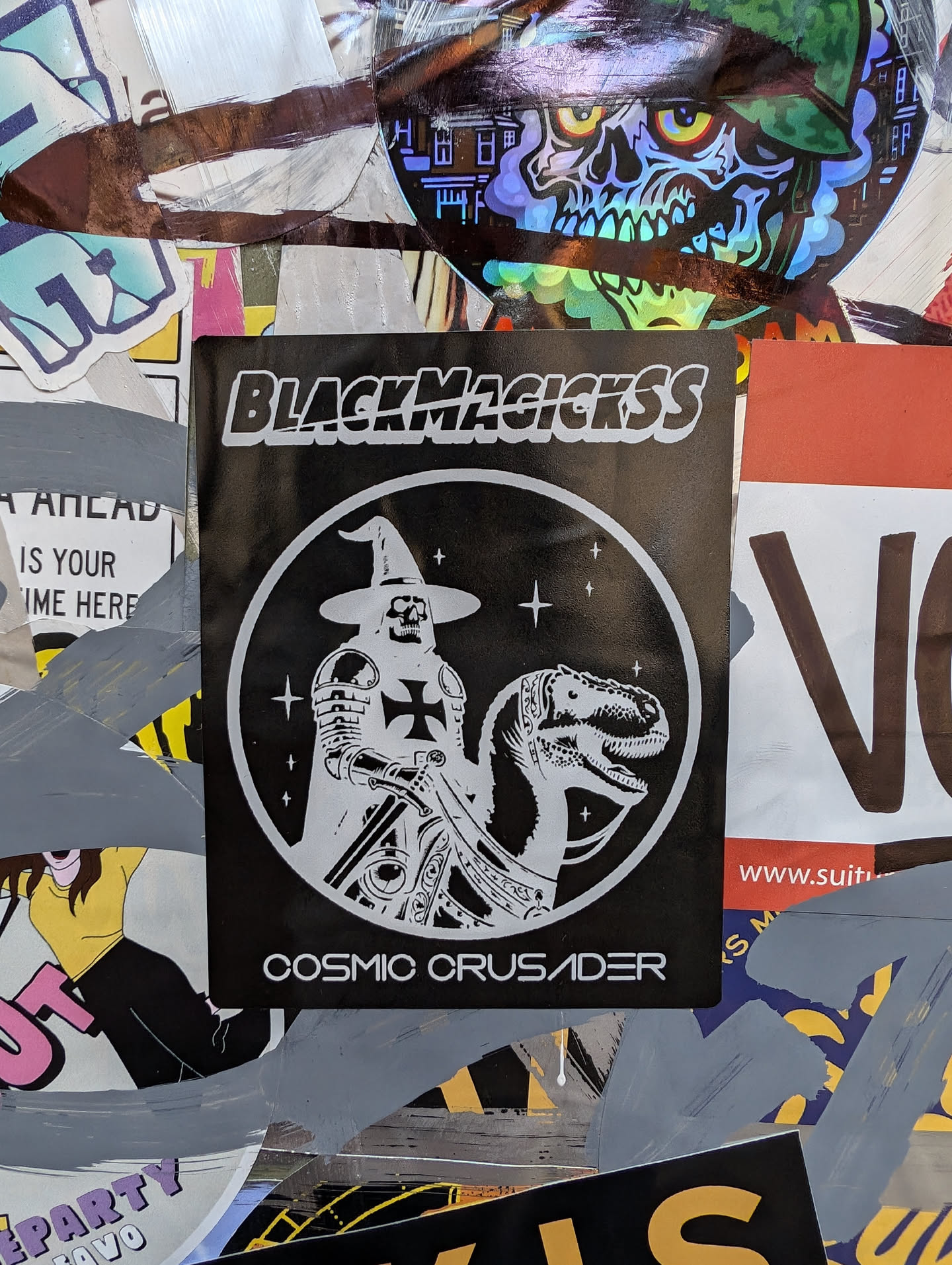 A black and white sticker featuring a knight riding a dinosaur, with the text "BlackMagickss" at the top and "Cosmic Crusader" at the bottom. The design is enclosed in a circle.