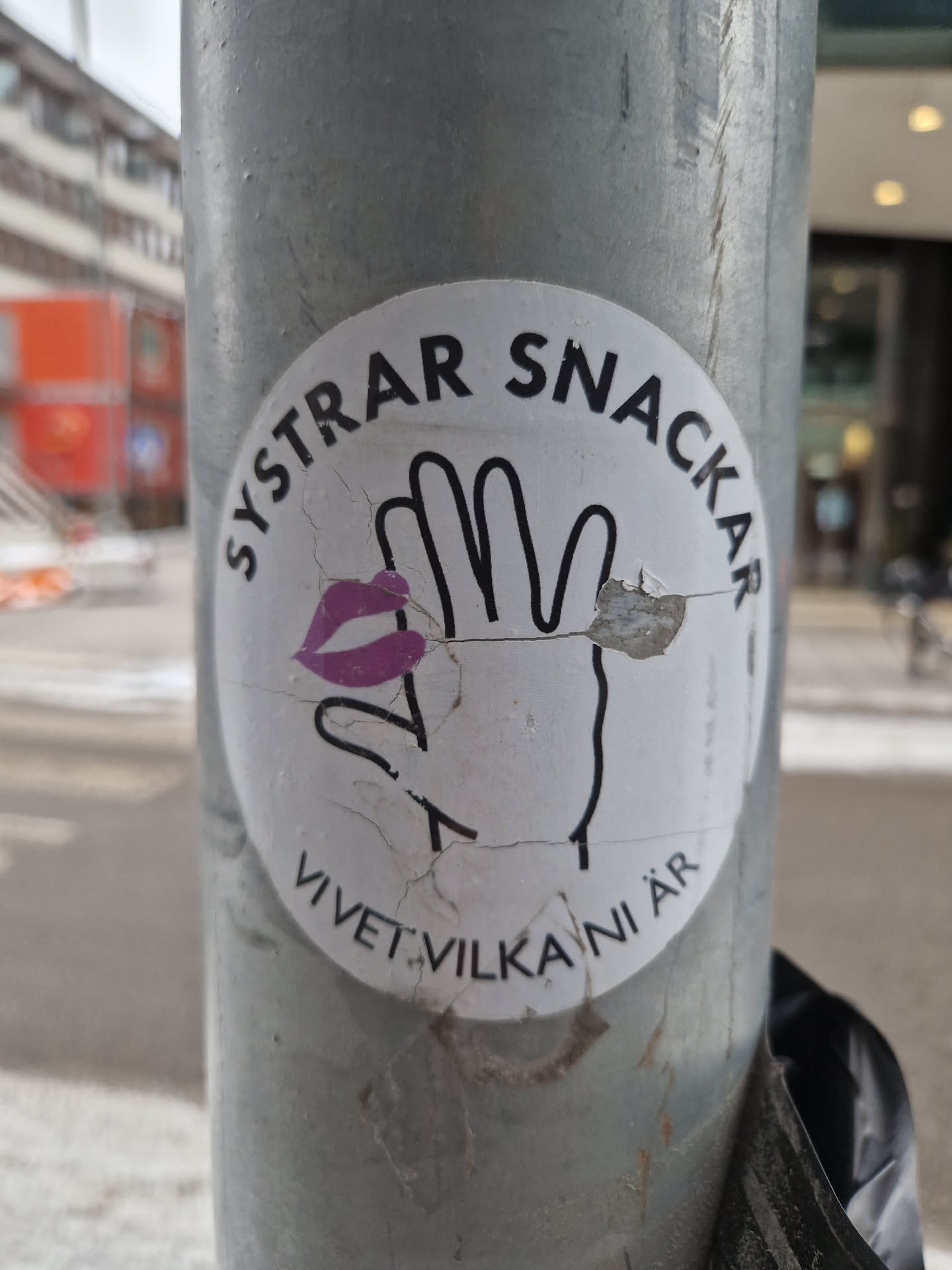 A round sticker featuring a hand with a purple lip print and the text "Systrar Snackar" around the top and "Vivet vilka ni är" around the bottom. The sticker is slightly damaged.