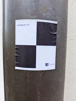 The image shows a rectangular sticker affixed to a dark, cylindrical object, possibly a pipe or pole. The sticker features a black and white checkered pattern, with four squares arranged in a 2x2 grid. The upper left corner of the sticker reads "Kontrollpunkt: 570". Below the checkered pattern, the logo of newCAD is visible. The sticker appears slightly wrinkled and weathered.