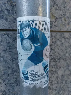 A weathered sticker featuring a retro illustration of a soccer player. The text on the sticker appears to be in Finnish.  The sticker is affixed to a metal pole near a City Hall. 