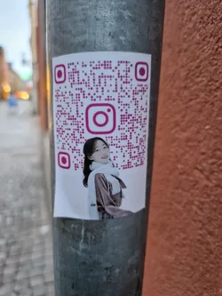 A sticker featuring a young Asian woman wearing winter clothes. The sticker includes a QR code linking to an Instagram account. The sticker is affixed to a metal post outdoors.