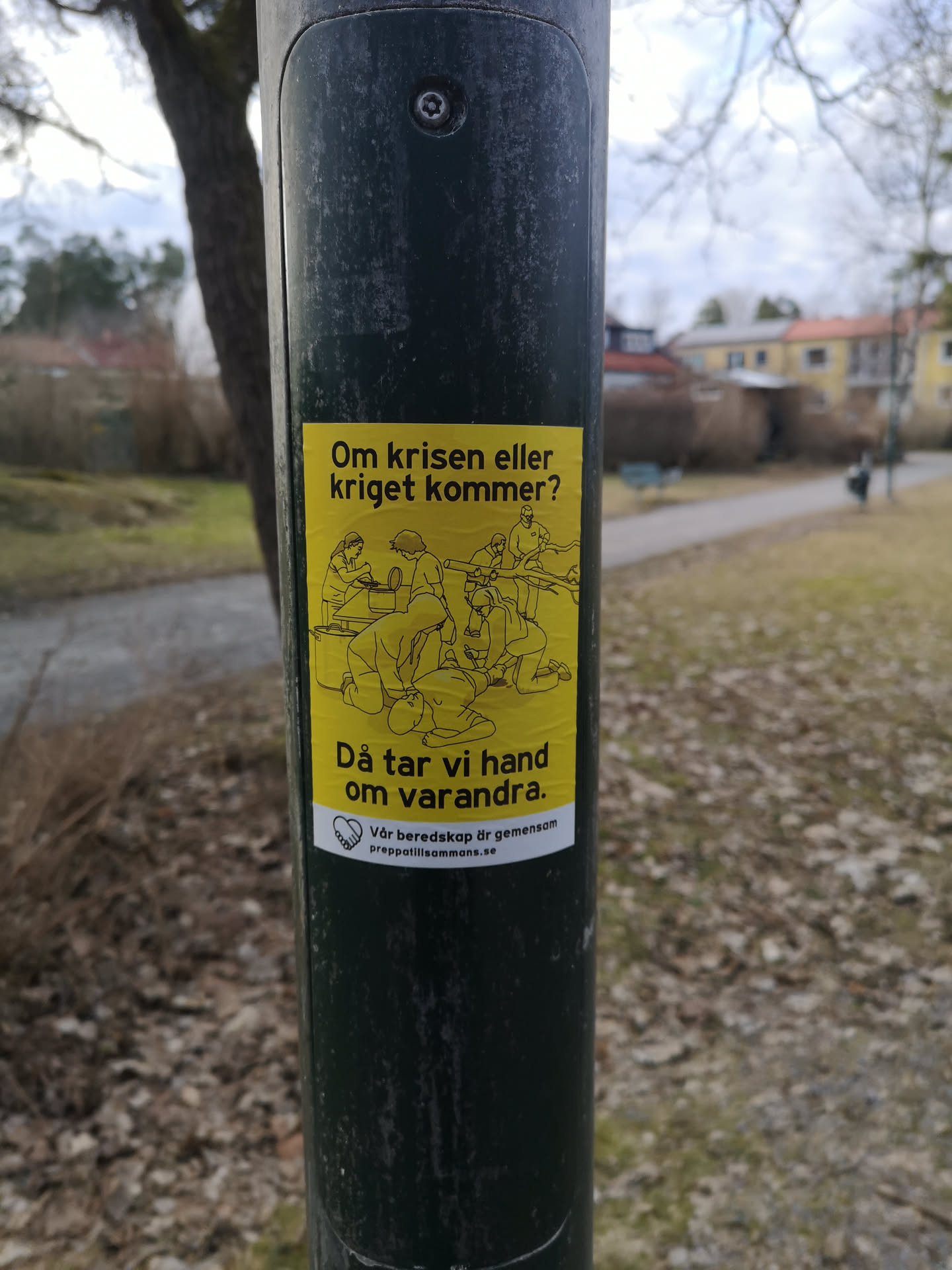 The sticker is rectangular with a vibrant yellow background and features black line art illustrations. At the top, the text "Om krisen eller kriget kommer?" is written. Below this, a group of approximately seven stylized figures are depicted in various acts of assistance and preparation, including tending to an injured person, cooking, and gathering resources like firewood. Below the illustration, the text "Då tar vi hand om varandra." is displayed. At the bottom, a small white heart icon containing two shaking hands is present, followed by the text "Vår beredskap är gemensam" and the website address "preppatillsammans.se". The overall aesthetic is simple, illustrative, and conveys a strong message of collective action and support.