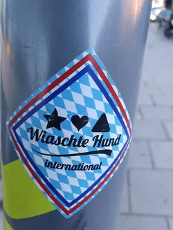 A diamond-shaped sticker with a Bavarian pattern features the text "Wiaschte Hund international." It includes three symbols: a star, a heart, and a triangle. 