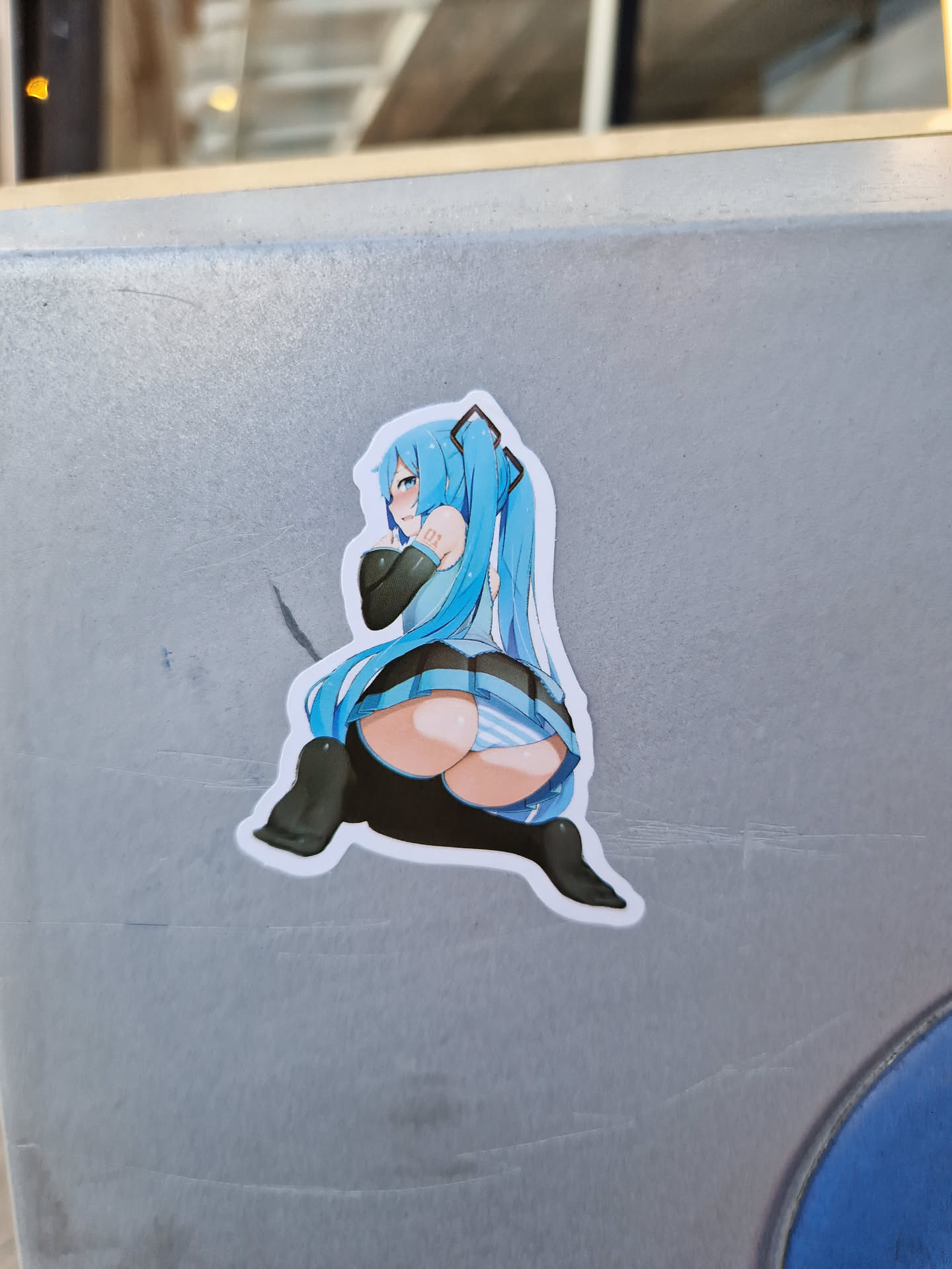 A vinyl sticker of an anime girl in a blue and white outfit. She is shown from behind, and her long, bright blue hair is visible. The sticker is affixed to a gray surface.