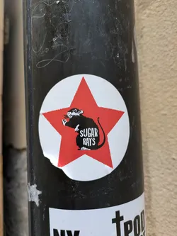 A round sticker with a red star and a black rat in the center. The text 'Sugar Rats' is written below the rat. The sticker is attached to a black pole.