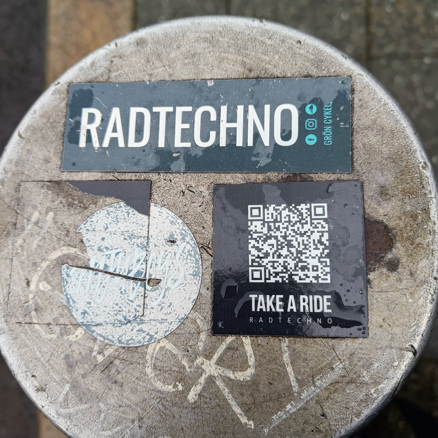 A black and white QR code sticker with the text "TAKE A RIDE" and the logo "RADTECHNO". There is also a larger sticker with the word RADTECHNO and social media links for Instagram and Facebook. The stickers are on a gray surface.