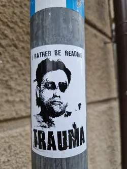 Black and white sticker featuring a portrait of a man with an eye patch, with text overlay above and below the image. The top text says "I rather be reading", and the bottom text says "Trauma".