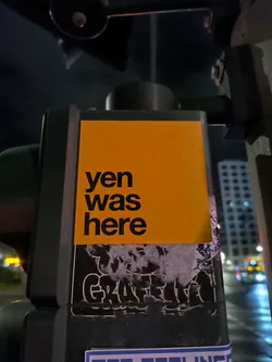 A yellow sticker with the text "yen was here" in black letters. It is attached to a dark gray surface, possibly a traffic light pole. There are other stickers visible underneath and around it.