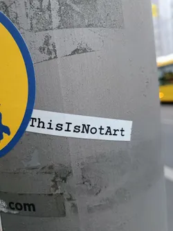 A minimalist sticker with the text "ThisIsN0tArt" printed on it. The sticker is affixed to a gray pole.