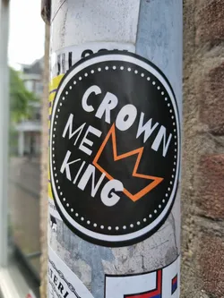 Round black and orange sticker with the text CROWN ME KING and a crown symbol in the center. The sticker is on a pole.