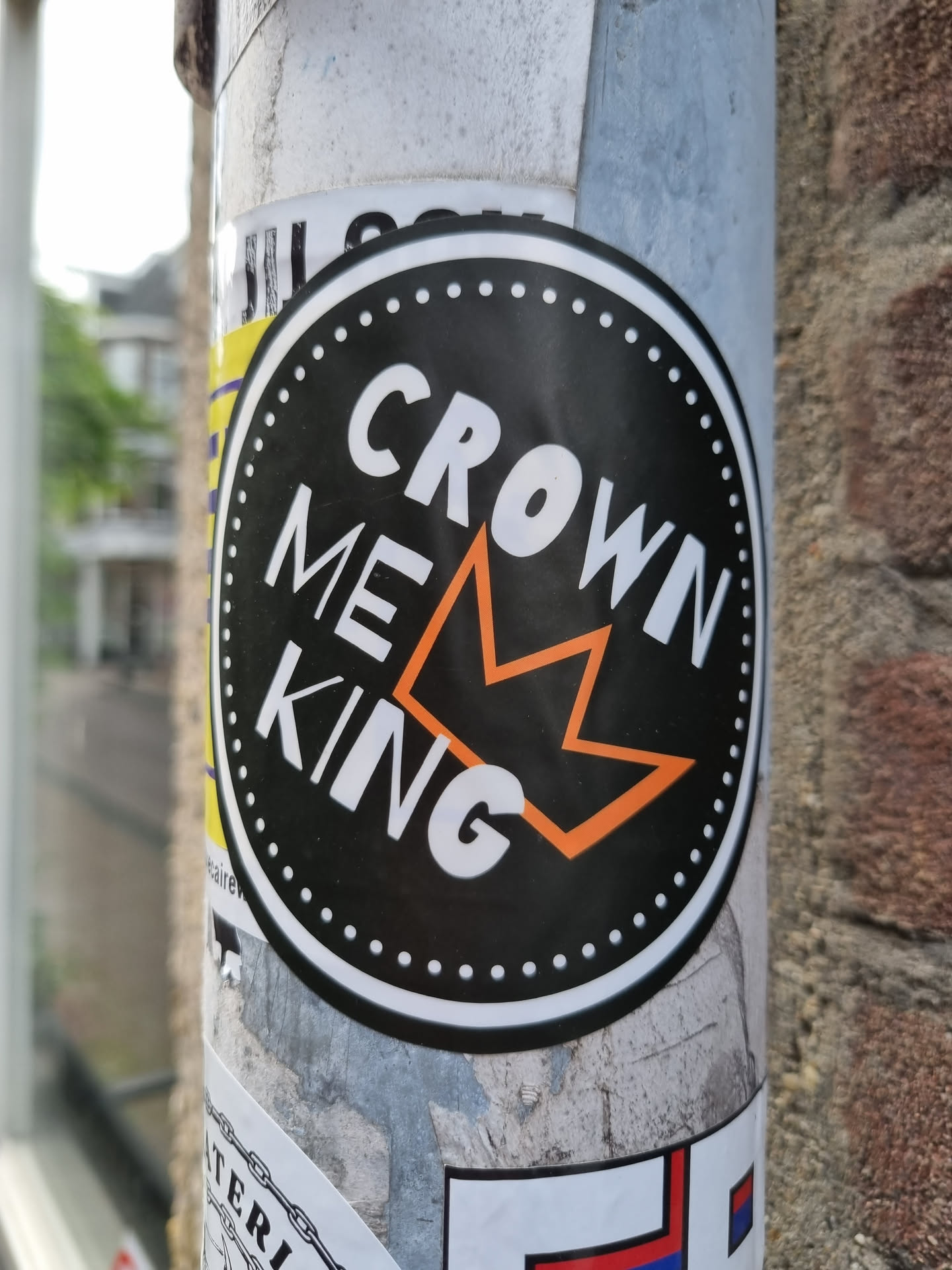 Round black and orange sticker with the text CROWN ME KING and a crown symbol in the center. The sticker is on a pole.