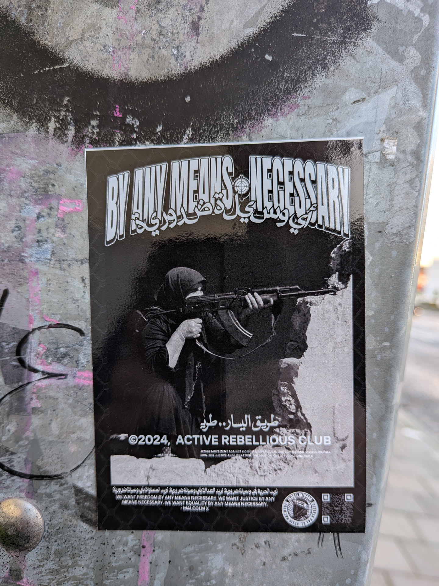 The sticker features a black and white image with a prominent title in bold, stylized English lettering that reads 'BY ANY MEANS NECESSARY'. Below this, in Arabic script, is text that appears to be a translation or related phrase. The central image depicts a woman wearing a hijab, holding and aiming an assault rifle. She is positioned behind a rough, textured surface that suggests cover or rubble. The background is dark, with a subtle mesh pattern. Below the image, the text '©2024, ACTIVE REBELLIOUS CLUB' is visible, along with smaller text that reads 'DIVIDE MOVEMENT AGAINST ZIONISM & IMPERIALISM, UNITED WE STAND, DIVIDED WE FALL. FIGHT FOR JUSTICE AND LIBERATION. ONE MISSION, ONE STRUGGLE, ONE FIGHT.' At the bottom, there is more text in Arabic and English, including the quote 'WE WANT FREEDOM BY ANY MEANS NECESSARY, WE WANT JUSTICE BY ANY MEANS NECESSARY, WE WANT EQUALITY BY ANY MEANS NECESSARY. - MALCOLM X'. A circular emblem and a QR code are also present on the lower right side.