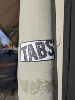 The sticker is rectangular with a white background and a thick black border. The word 'TABS' is printed in large, bold, black sans-serif capital letters. The sticker appears slightly worn, with some scuff marks and imperfections on its surface and edges, suggesting it has been exposed to the elements.