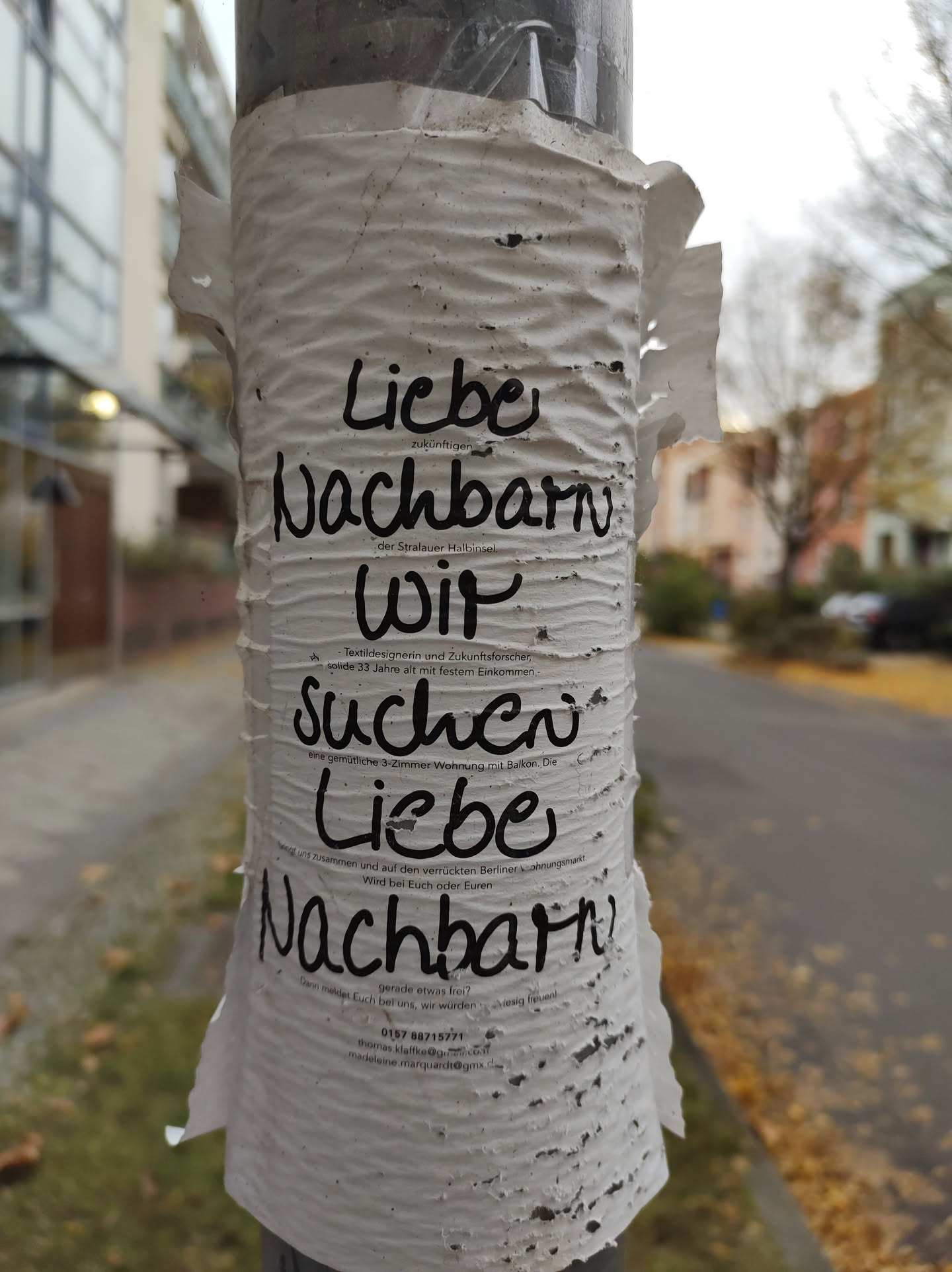 A worn-out sticker on a pole with a handwritten message in German. The message says: "Liebe Nachbarn wir suchen liebe Nachbarn", which translates to: "Dear neighbors we are looking for dear neighbors".