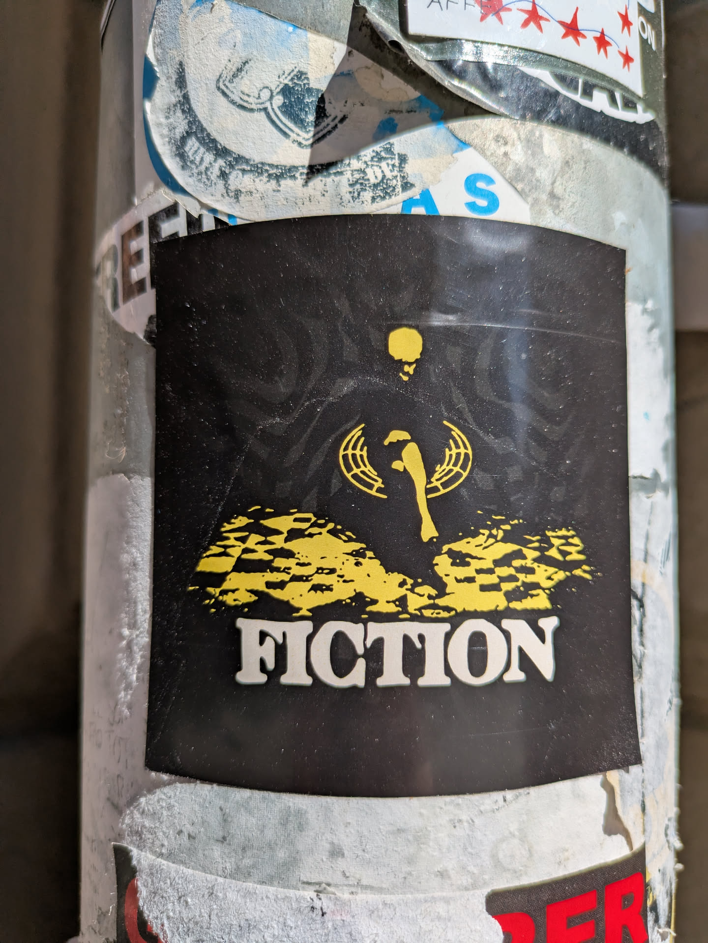 The sticker has a black background with a yellow figure above the word "FICTION". The figure appears to be a person or skeleton holding a scepter, surrounded by a geometric design. The word "FICTION" is printed in a bold, white font.