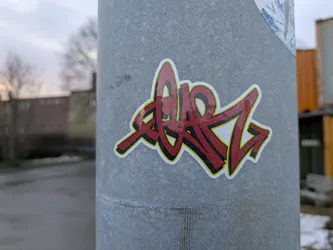 A sticker with red graffiti art on a gray metal pole. The graffiti is stylized and dynamic, with sharp angles and curves. The background of the sticker is yellow, providing a contrast to the red graffiti. The pole is textured and shows signs of wear.