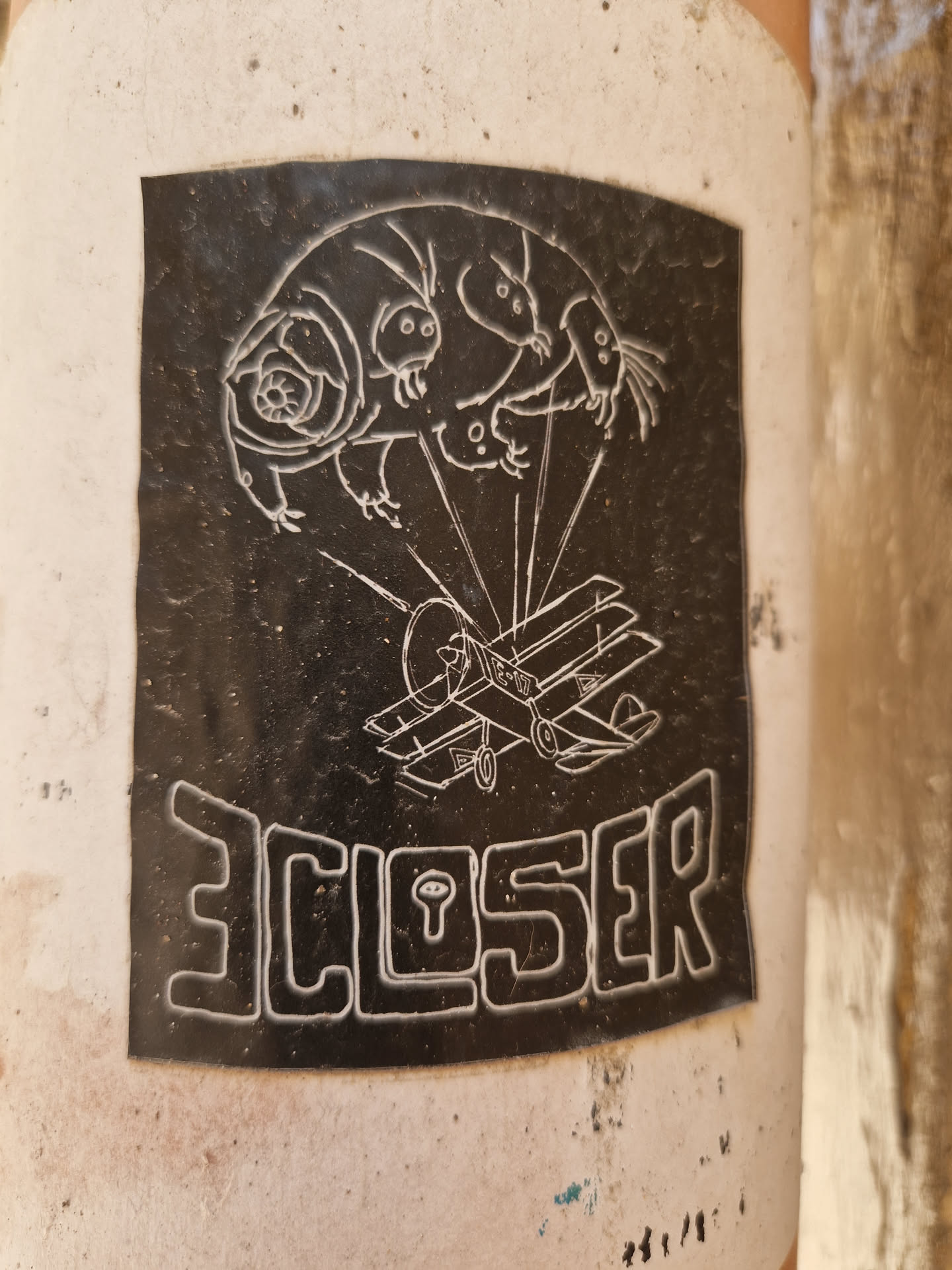 Black and white sticker featuring a stylized illustration of a tardigrade seemingly piloting a small biplane. The word 'CLOSER' is written in a stylized font at the bottom. The image is slightly worn, suggesting it's been affixed outdoors for some time.