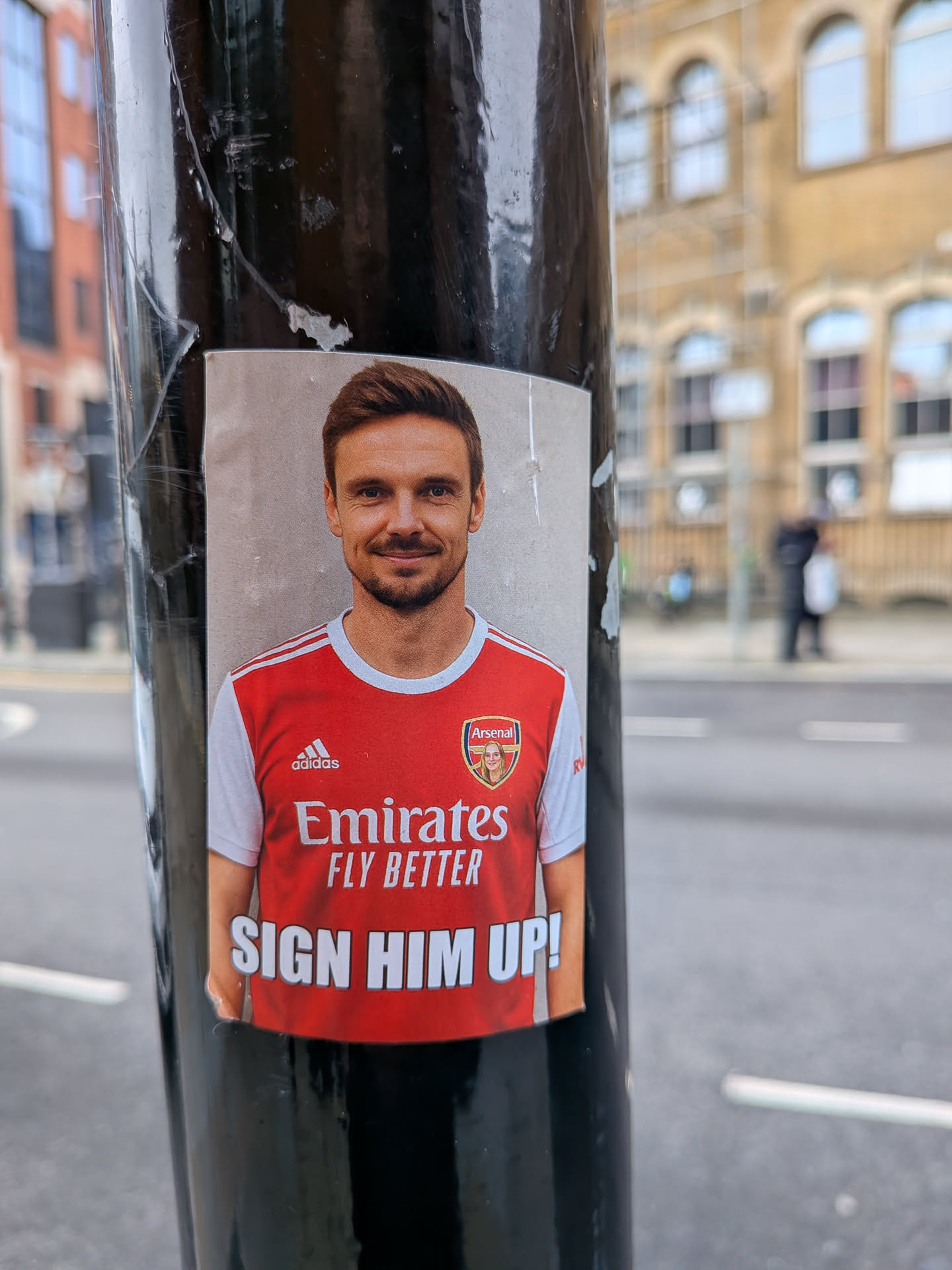 A rectangular, vertically oriented sticker featuring a smiling man with short brown hair and a light beard, wearing a red and white Arsenal home jersey. The Adidas logo is visible on the right chest, and the Arsenal crest is on the left. Uniquely, the traditional cannon within the Arsenal crest has been replaced with a small, circular photo of a blonde woman. Below the crest, the main sponsor 'Emirates FLY BETTER' is displayed in white text. At the bottom of the sticker, in large white capital letters on a red background, is the phrase 'SIGN HIM UP!'. The background behind the man is a plain, light grey or beige color, suggesting a wall. The sticker is adhered to a black pole, showing some minor creases and wear around its edges.