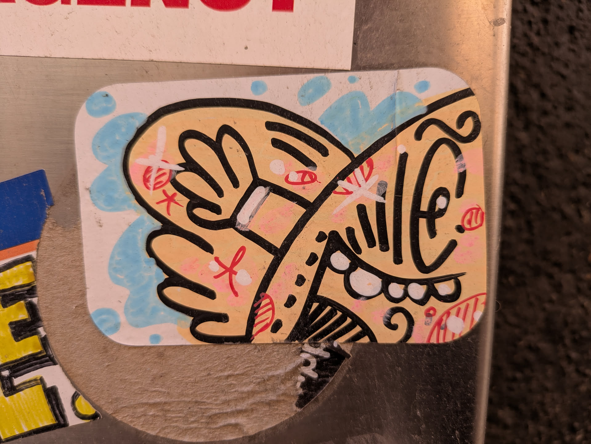 A cartoon face sticker with a light tan base color and black outlines. The face is framed by two hands in a similar style. The background has light blue accents resembling water droplets. The artwork uses pink and red for additional accents. The face features exaggerated features such as a large eye, prominent teeth, and other whimsical details.