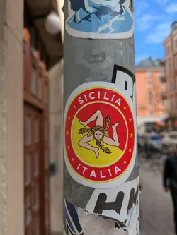 A round sticker affixed to a gray pole features a red border with yellow stars, encircling a yellow interior. Centered within the yellow area is an emblem of Trinacria, a head with three legs emanating from it. The sticker is labelled 'SICILIA' at the top and 'ITALIA' at the bottom.