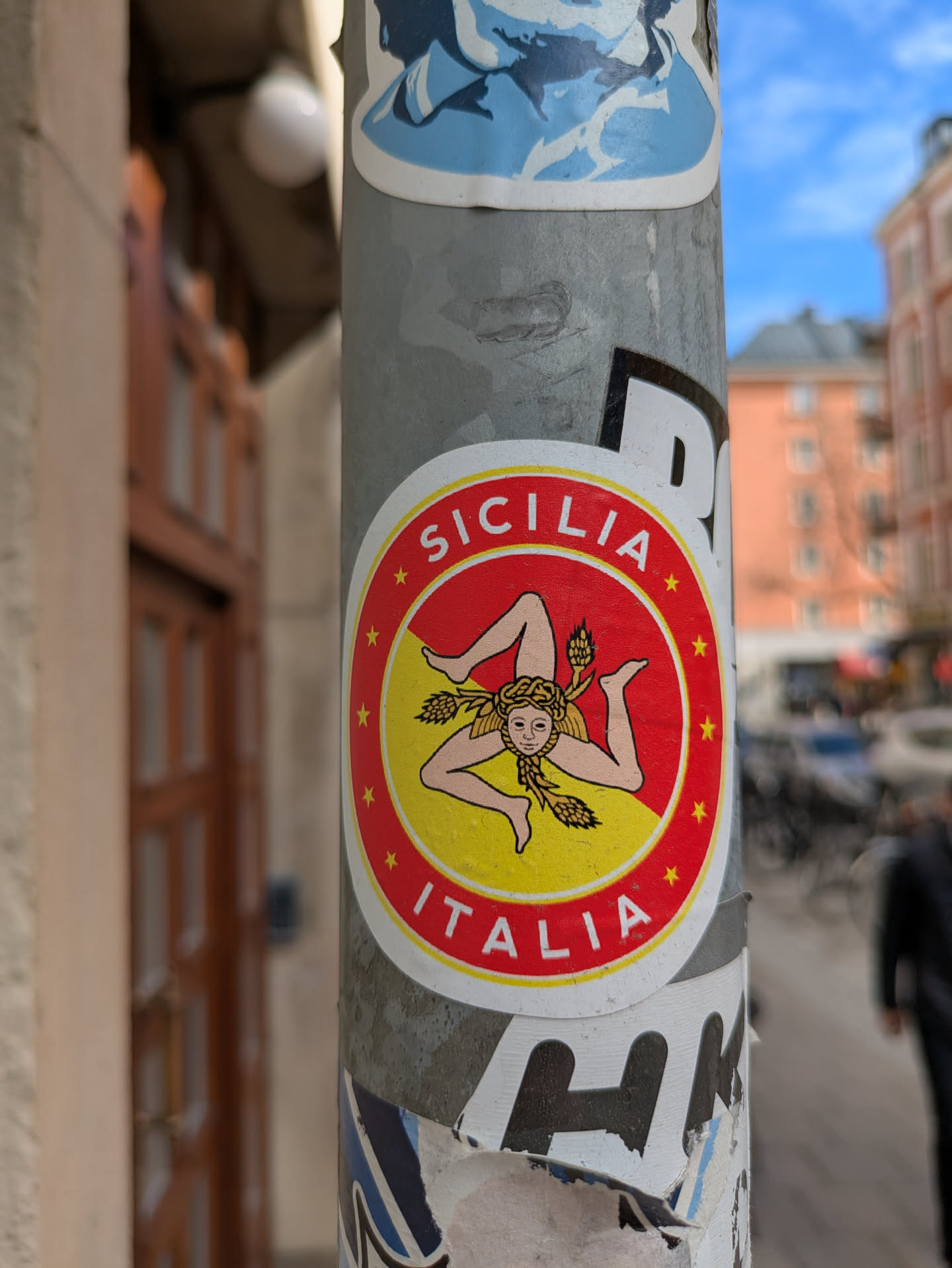 A round sticker affixed to a gray pole features a red border with yellow stars, encircling a yellow interior. Centered within the yellow area is an emblem of Trinacria, a head with three legs emanating from it. The sticker is labelled 'SICILIA' at the top and 'ITALIA' at the bottom.