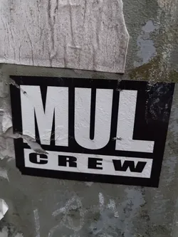 Black and white sticker with the text MUL CREW. The sticker is slightly damaged and is attached to a weathered surface.