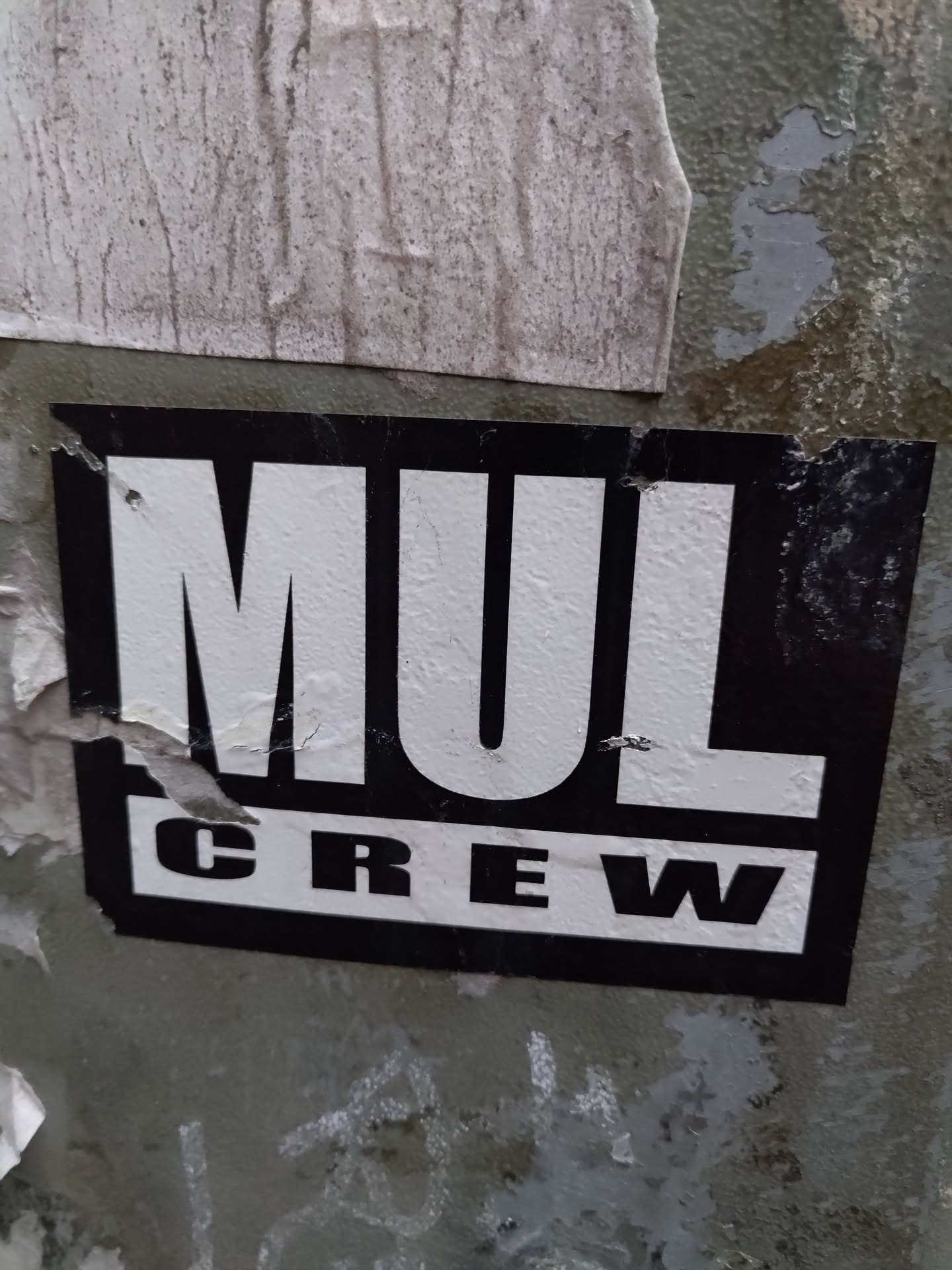Black and white sticker with the text MUL CREW. The sticker is slightly damaged and is attached to a weathered surface.