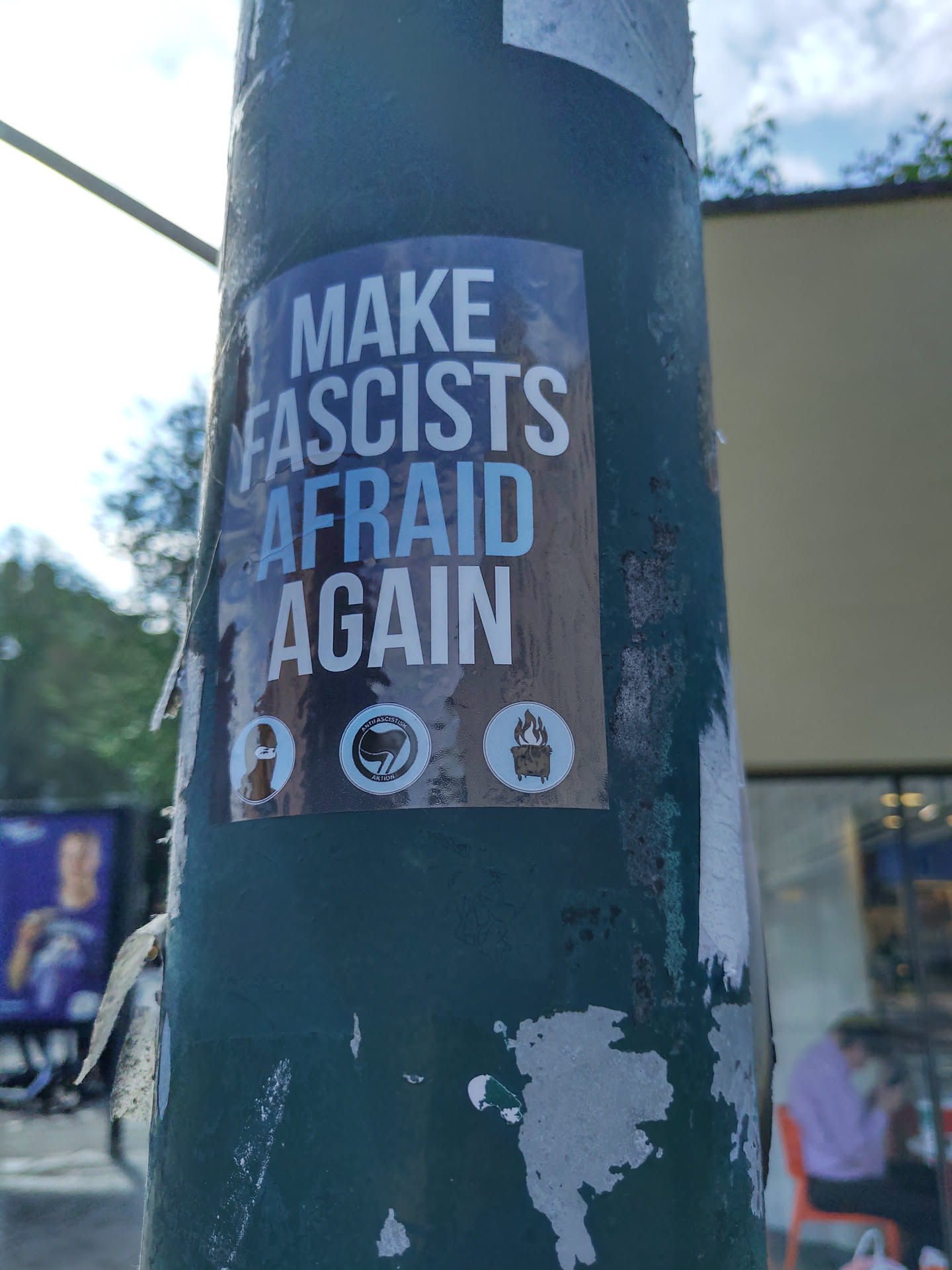 A sticker featuring the text "Make Fascists Afraid Again" and three symbols. The sticker is affixed to a pole.