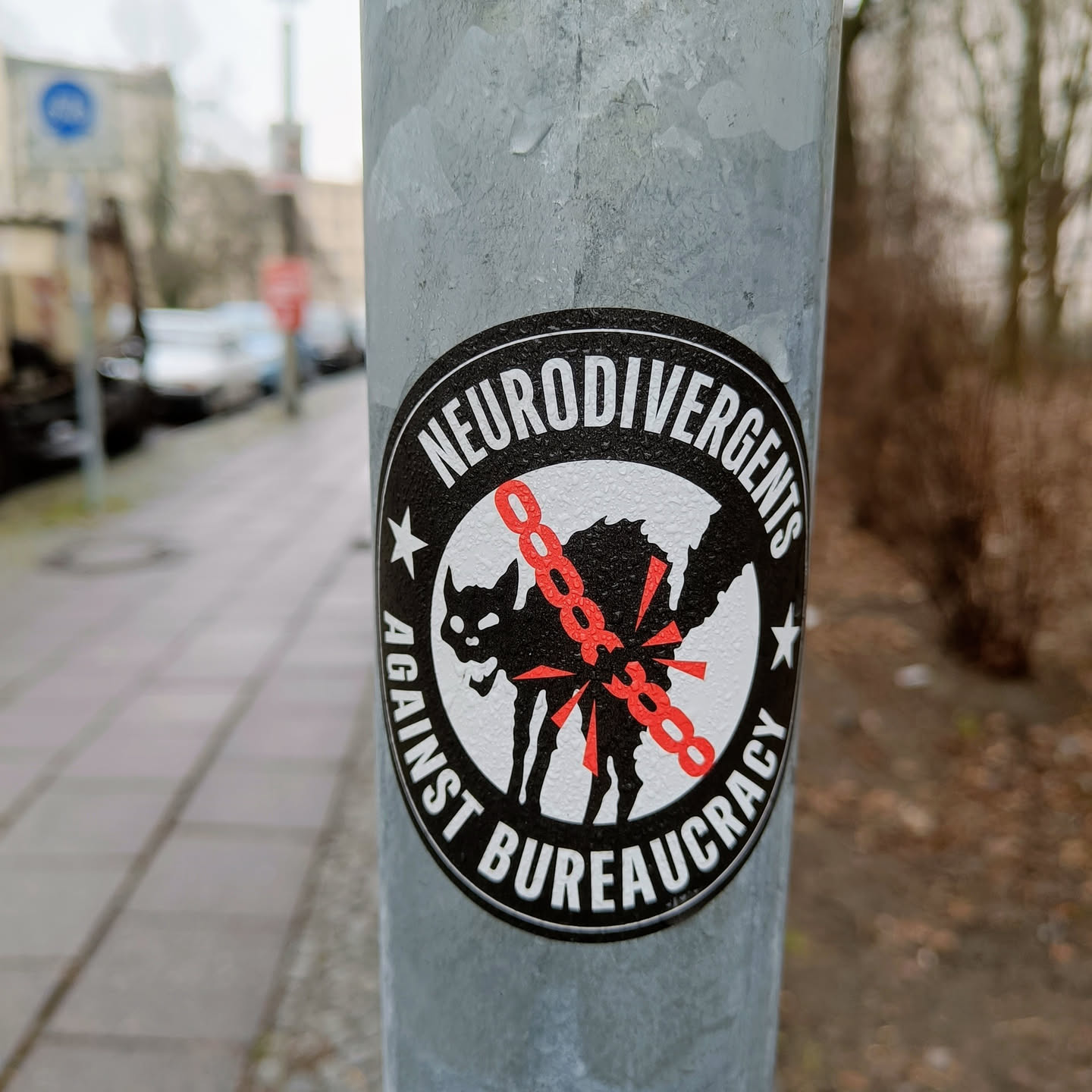 A black and white sticker with a black cat breaking free from red chains. The text 'Neurodivergents Against Bureaucracy' is written around the cat.