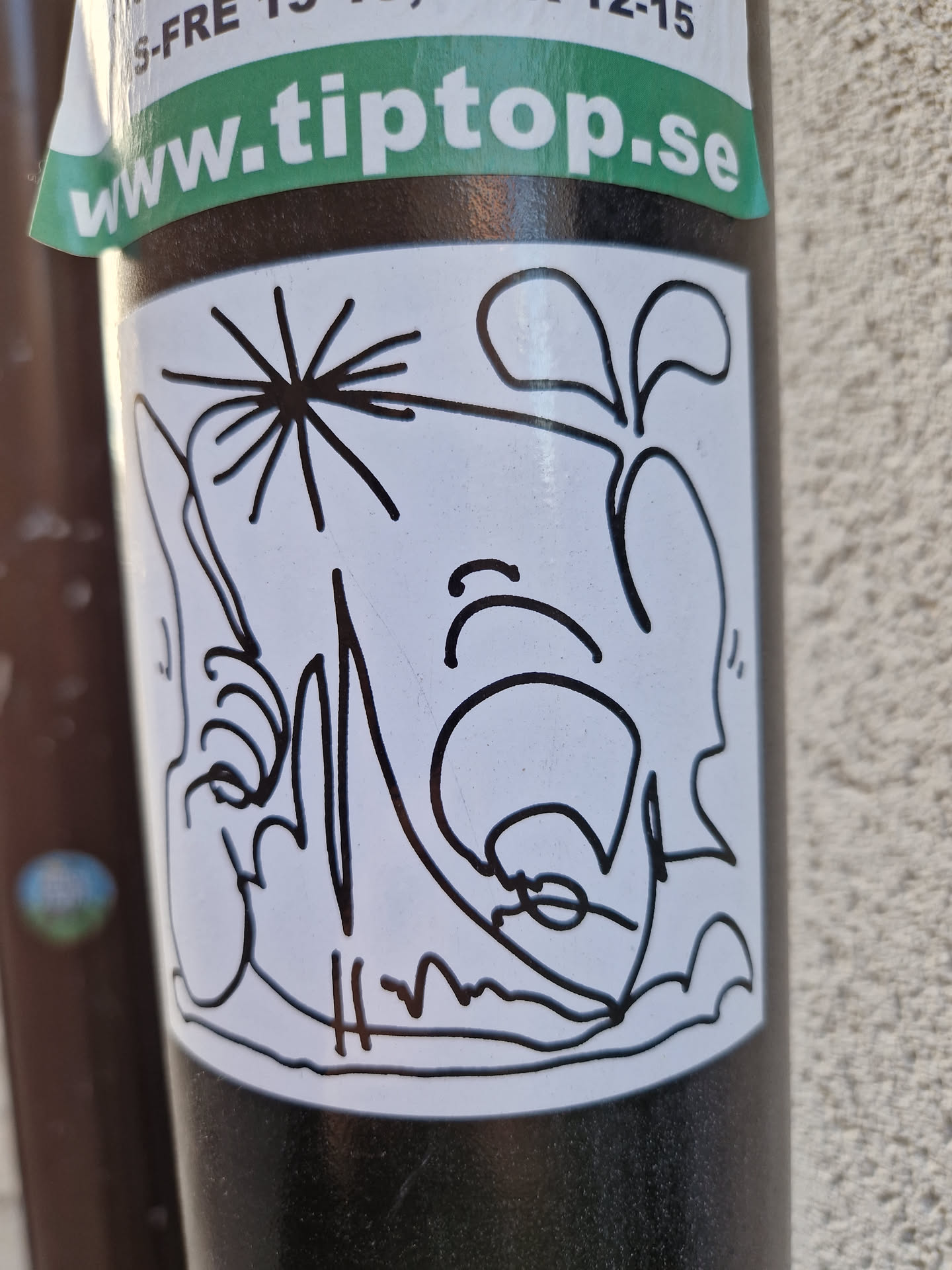A white sticker with a black ink drawing of a character that looks like a combination of a face and a plant. The style is simple and expressive, with visible brushstrokes giving a hand-drawn quality. There is a small signature-like mark at the bottom.