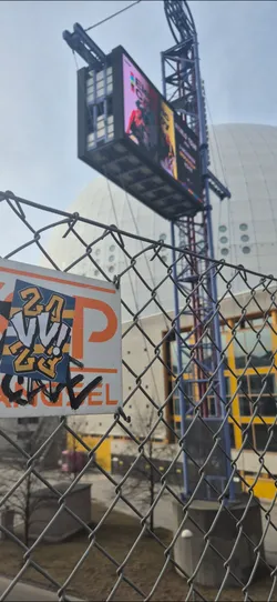 A close-up shot captures a rectangular sticker affixed to a chain-link fence. The sticker features the text "24 VI 2023" in bold letters and the name "GRRRL" written beneath. The colors of the text and background of the sticker are a mix of orange, white, and blue. The fence is a gray chain-link construction. The backdrop features a large, spherical building under a cloudy sky.