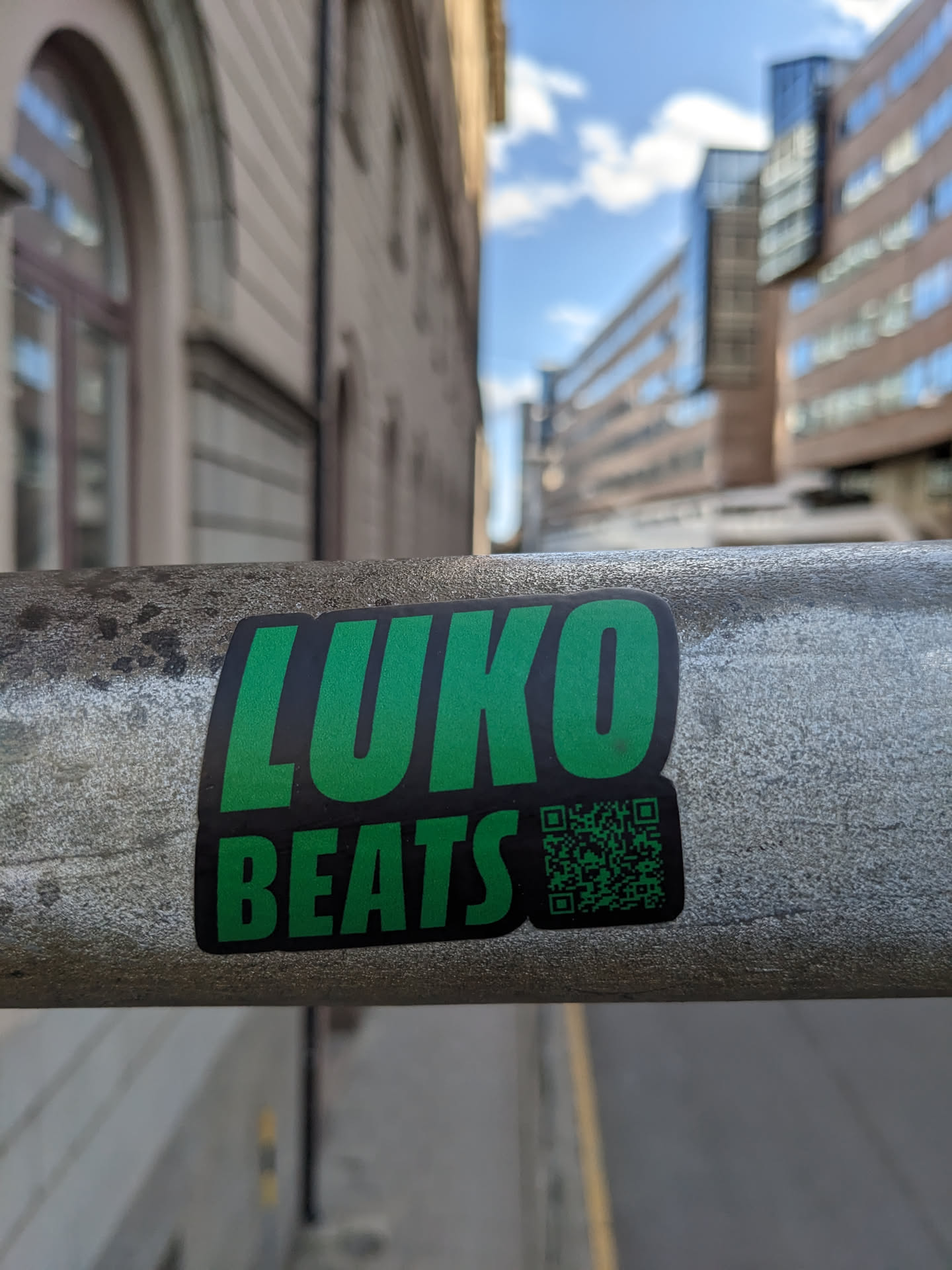 A green and black sticker with the text "LUKO BEATS" and a QR code. The sticker is affixed to a metal railing in an urban setting.