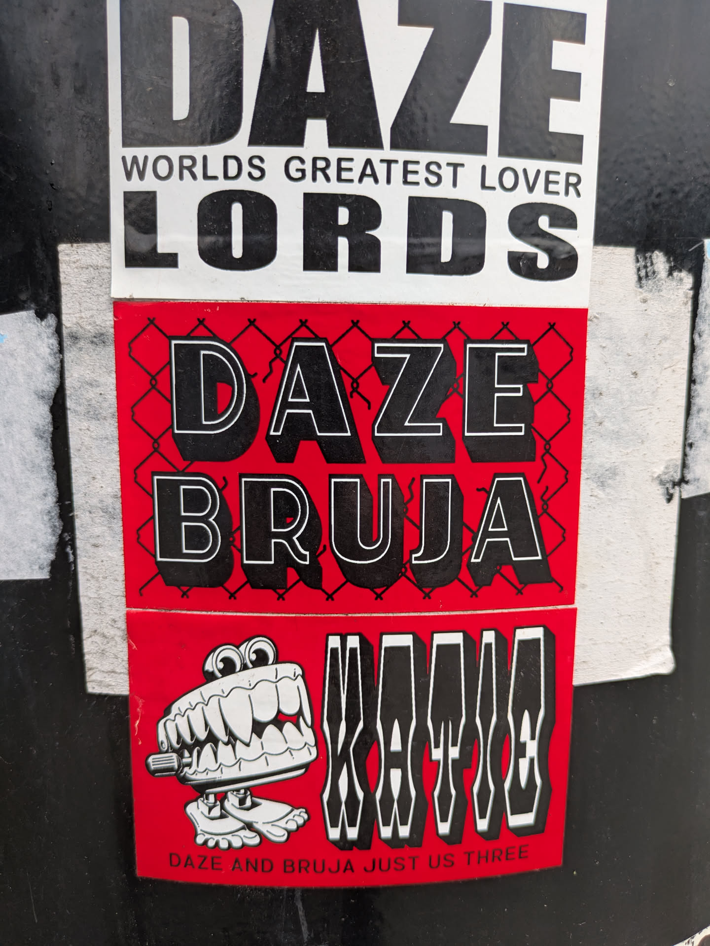 This multi-part sticker is predominantly red and black. The central section displays the words "DAZE BRUJA" in large, black font with a white outline, set against a red background with a black chain-link fence pattern. Below this, another red section features a whimsical illustration of wind-up chattering teeth with prominent eyeballs, positioned next to the word "KATTE" rendered in a striking, distorted black and white font. At the very bottom, in smaller white text, it reads "DAZE AND BRUJA JUST US THREE."