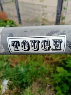 A sticker with the word TOUCH in a vintage style font. The sticker is attached to a grey metal pole. The background is blurred, showing some greenery and a metal fence.