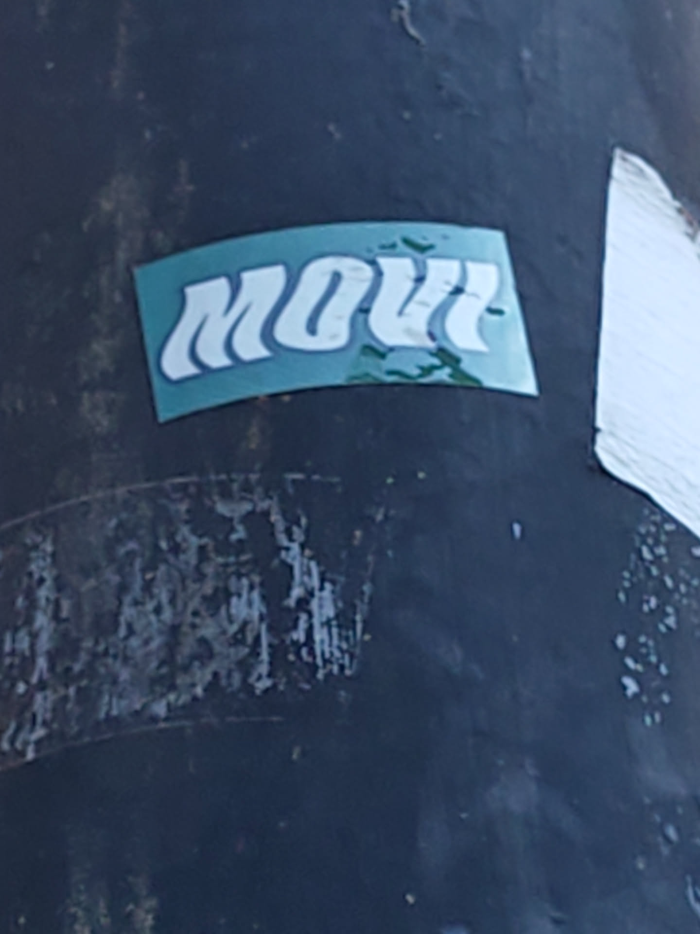 A teal and white sticker with the word "MOVI" in a stylized font. The sticker is affixed to a dark-colored pole or surface.