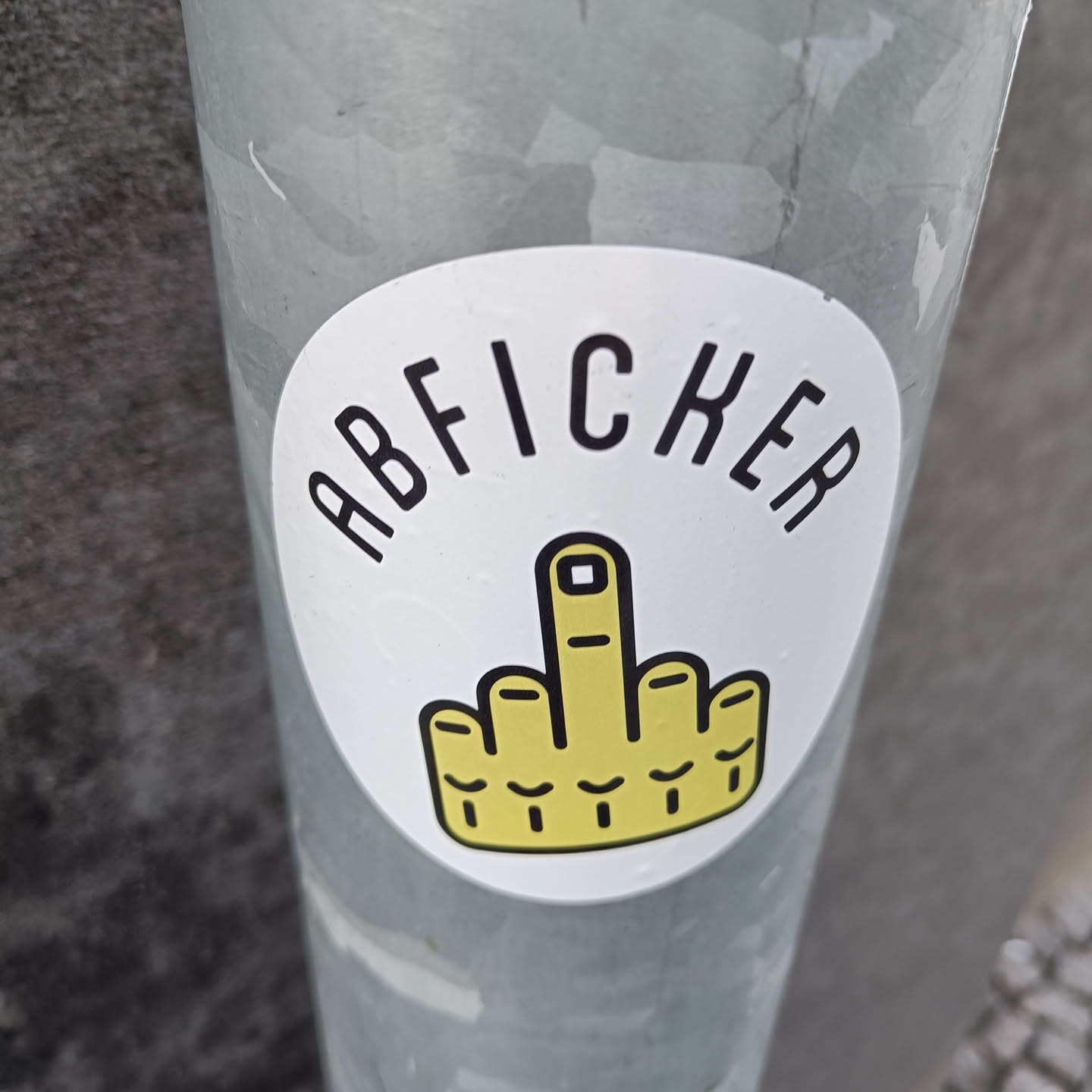 A round sticker with the word "ABFICKER" and a middle finger in yellow. The sticker is on a grey metal pole.