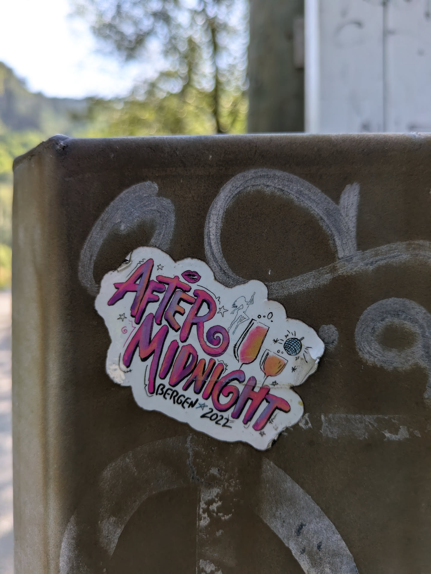 The sticker showcases the text 'After MIDNIGHT' in a bold, graffiti-style font with a pink and purple gradient. Above the text, there are playful illustrations including a pair of lips, a dancing figure, two wine glasses, and a sparkling disco ball, all hinting at a party or nightlife theme. Below the main phrase, 'BERGEN ★ 2022' is written, indicating a specific location and year, likely for an event. The sticker itself has a distinctive wavy, cloud-like white border and is placed on a dark, textured surface, possibly a utility box or wall, which also has some white graffiti marks.