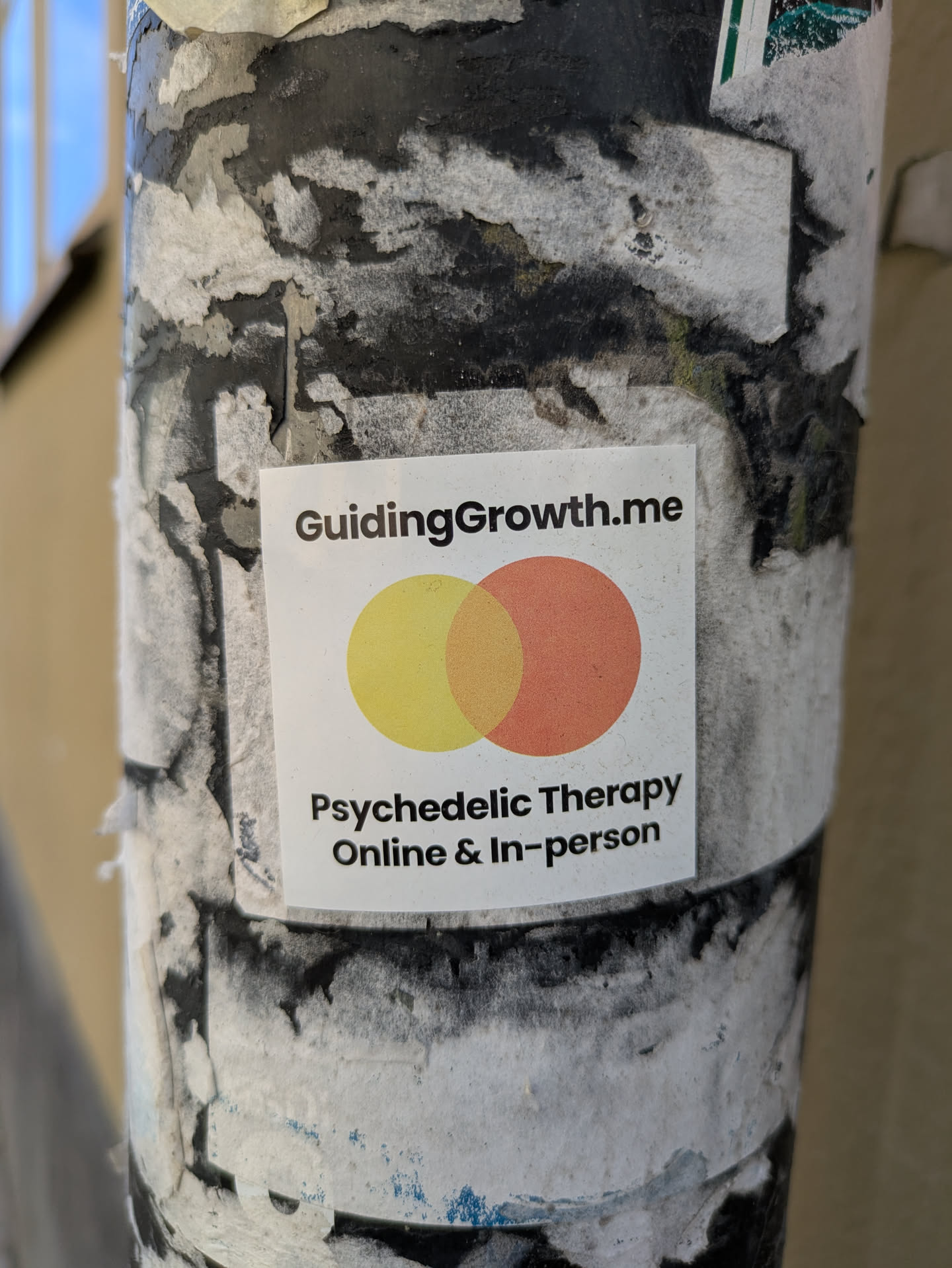 A square sticker is adhered to a post covered in peeling paper. The sticker has the text 'GuidingGrowth.me' at the top, followed by two overlapping circles, one yellow and one peach. Below the circles, the text 'Psychedelic Therapy Online & In-person' is printed.