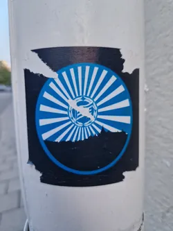 A partially peeled sticker featuring a circular design. The center contains a stylized sun with rays emanating outwards, overlaid on a dark silhouette of a cityscape. The color scheme is primarily blue and black.