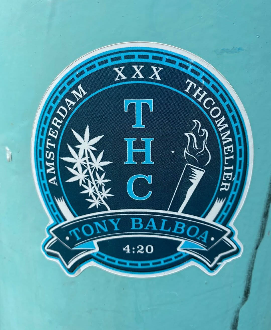 Round sticker with dark blue background and light blue accents. It features the text 'AMSTERDAM' and 'THCOMMELIER' arc-shaped at the top and bottom, with 'XXX' at the top center. In the center, it says 'THC' in large letters, with a marijuana leaf image to the left and a lit joint to the right. At the bottom, it says '.TONY BALBOA.' with '4:20' below. 