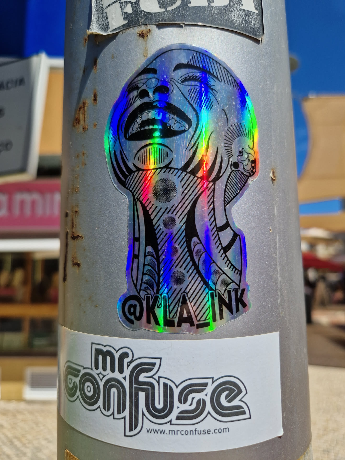 This iridescent sticker features a striking image of a laughing person's face, rendered in a detailed, almost geometric style.  The holographic effect shifts colors depending on the light. A smaller sticker underneath it displays "mr confuse" and a website address.