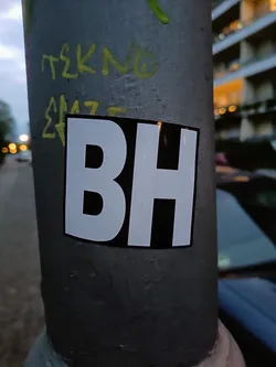 A simple yet striking black and white sticker with the letters BH in bold, uppercase font.  It's affixed to a grey metal pole, amongst other markings.