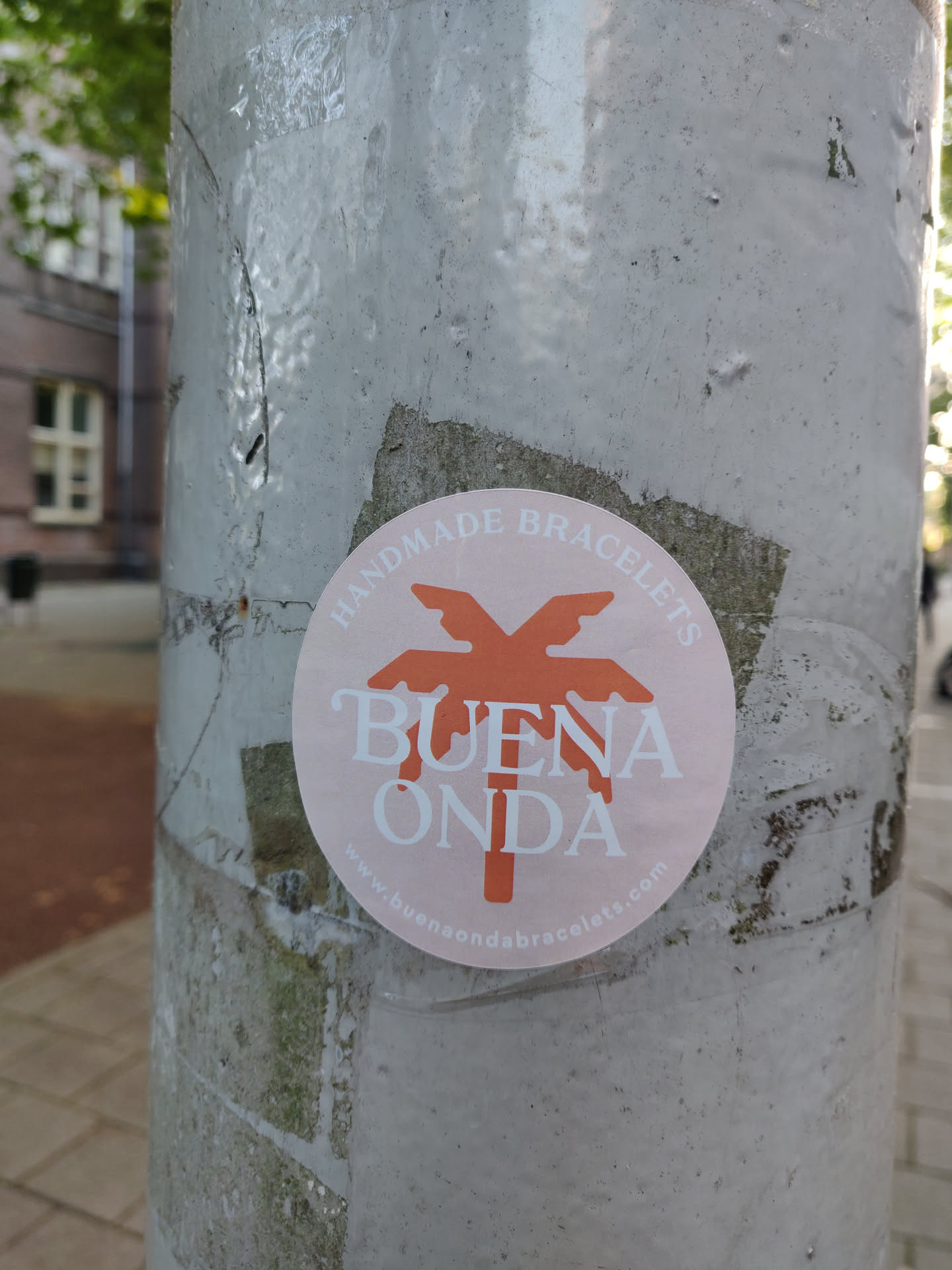 A round sticker with a palm tree design and the text 'BUENA ONDA' in the center. The text 'HANDMADE BRACELETS' is written around the circle. A website address is also included at the bottom of the sticker. The sticker is affixed to a gray pole.