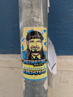 A pixelated sticker is affixed to a metal pole. The sticker features a pixel art portrait of a man with a mullet and a slight smile. Below the portrait, the text "Retratos 8bits" is displayed in pixelated font, followed by "+ whatsapp + 635347649" in a similar style. The background of the sticker is yellow with blue borders.