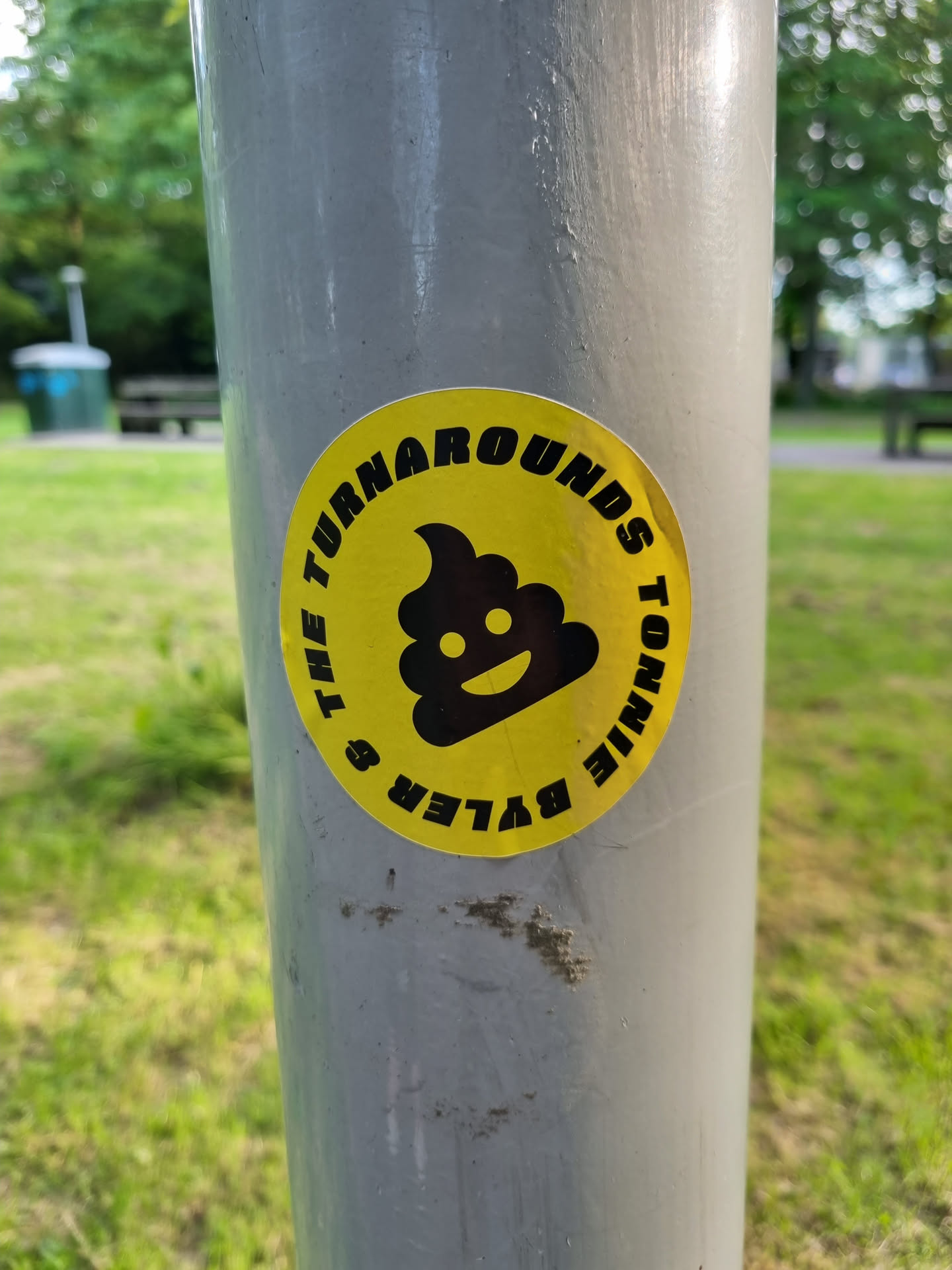 A yellow circular sticker with a black smiling poop emoji in the center and text surrounding it. The text reads: "Turnarounds Tonnie & The Byler's". The sticker is affixed to a grey metal pole.