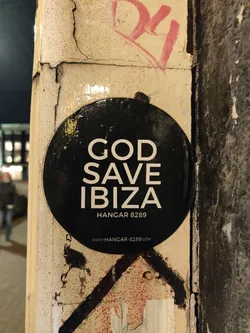 A black round sticker with the text "GOD SAVE IBIZA" in white uppercase letters. Underneath, it says "HANGAR 8289" and a website address: www.hangar-8289.com. The sticker is affixed to a weathered, graffiti-covered surface.