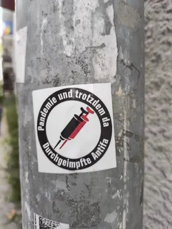 A sticker depicting two syringes with a circular text around it saying "Pandemie und trotzdem da Durchgeimpfte Antifa". It means Pandemic and still here vaccinated Antifa.