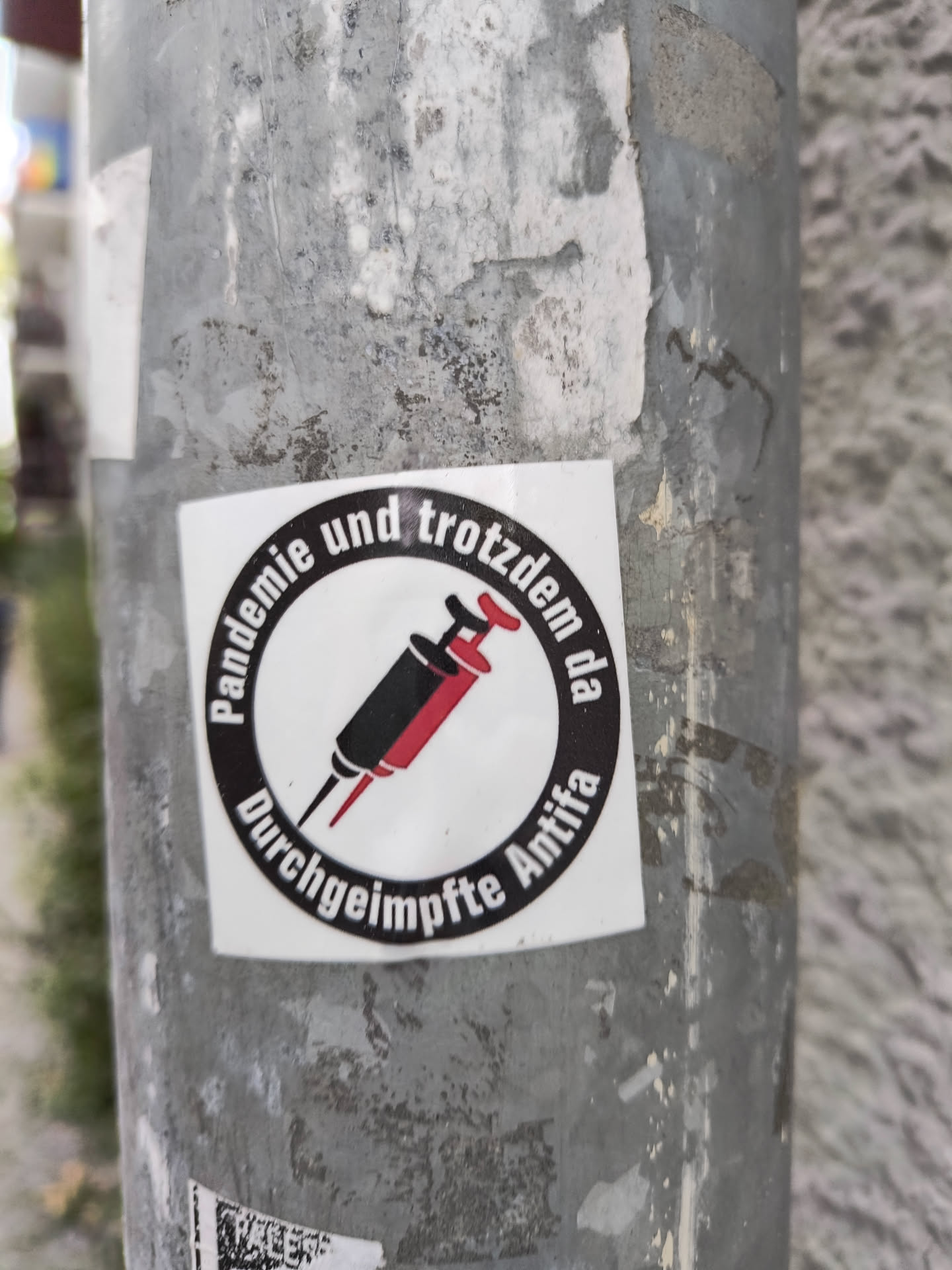 A sticker depicting two syringes with a circular text around it saying "Pandemie und trotzdem da Durchgeimpfte Antifa". It means Pandemic and still here vaccinated Antifa.