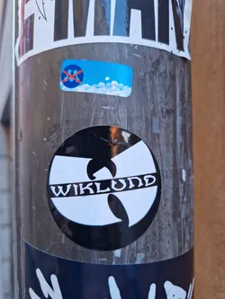 A round black and white sticker featuring a Wu-Tang Clan logo variation with the name "Wiklund" inscribed inside. The sticker is affixed to a pole, along with other stickers.