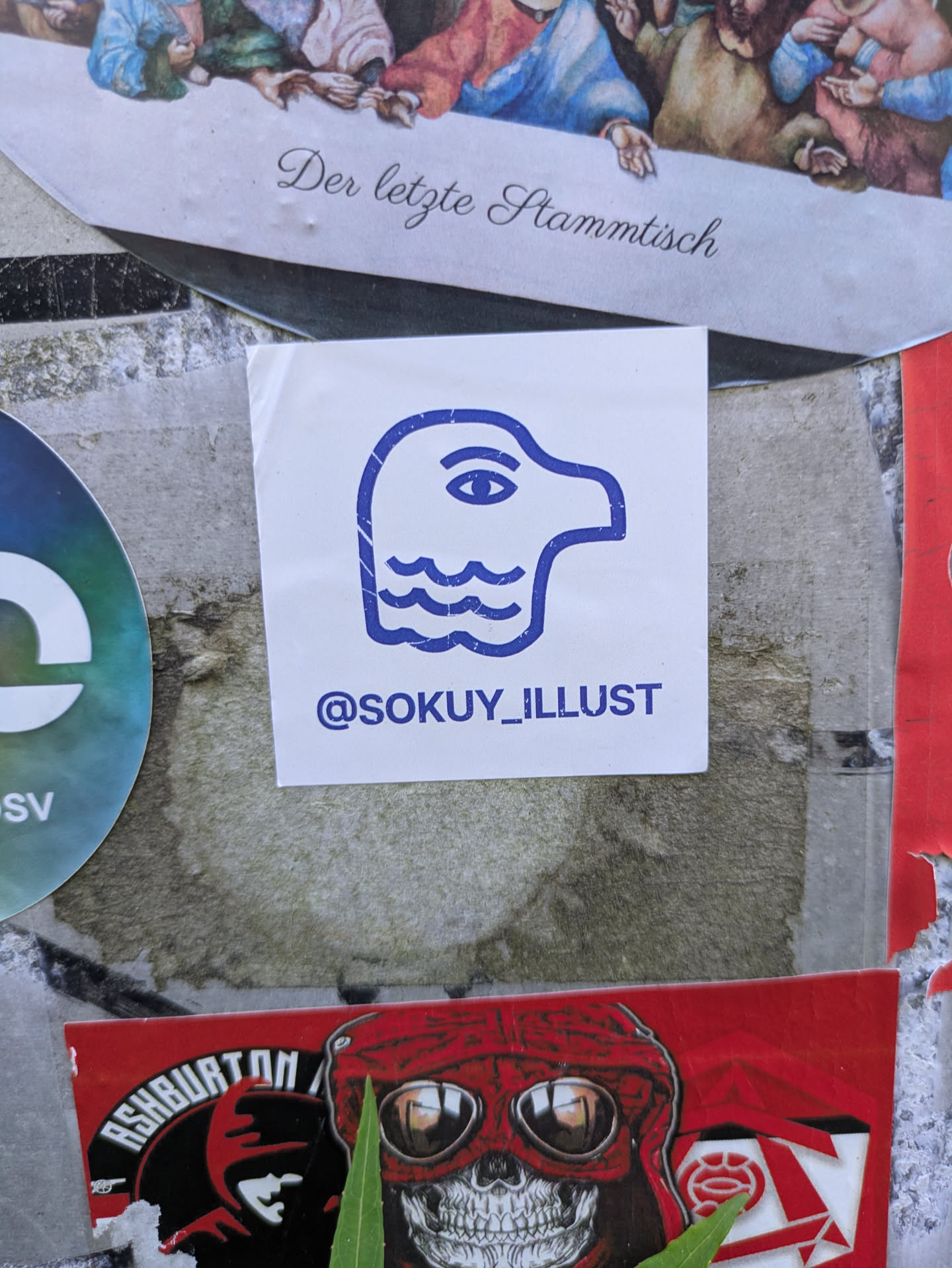 This central square white sticker features a distinct blue outline illustration. The image portrays a stylized, almost abstract head or profile, characterized by a prominent single eye and three wavy lines below it, suggesting water, sound, or a unique texture. The minimalist design gives it a modern aesthetic. Directly beneath the illustration, the text '@SOKUY_ILLUST' is printed in blue, likely serving as a creative alias or social media handle for an artist or illustrator. The sticker is affixed to a textured, possibly concrete or metal, surface, surrounded by other diverse stickers.